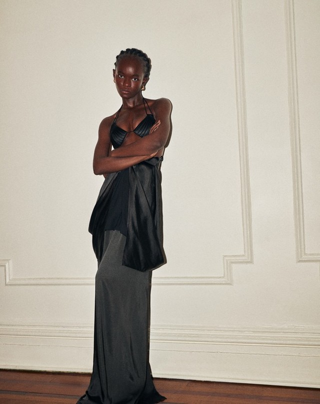 Full-length shot of a Black woman in a black bikini top and black and grey dress, arms crossed, against a plain wall.