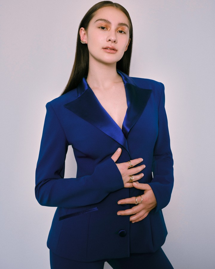 Adult female posing indoors wearing blue blazer and pants, accessorized with gold rings, against a light backdrop in portrait orientation.