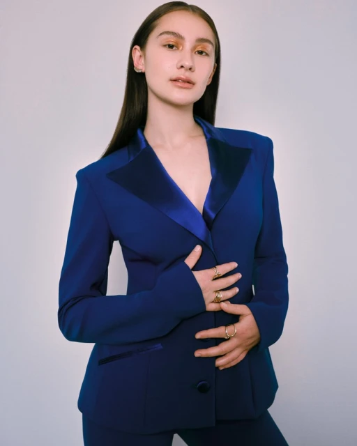 Adult female posing indoors wearing blue blazer and pants, accessorized with gold rings, against a light backdrop in portrait orientation.