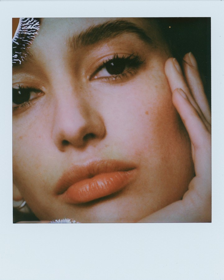 Close-up portrait of a young adult woman's face with dark eyes and full lips, touching her cheek with her fingers in a photo studio.