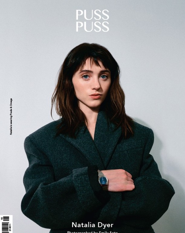 Portrait of Natalia Dyer wearing Prada and Omega on a magazine cover, medium shot on light grey studio background.
