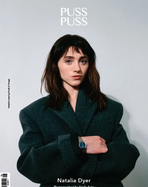 Portrait of Natalia Dyer wearing Prada and Omega on a magazine cover, medium shot on light grey studio background.