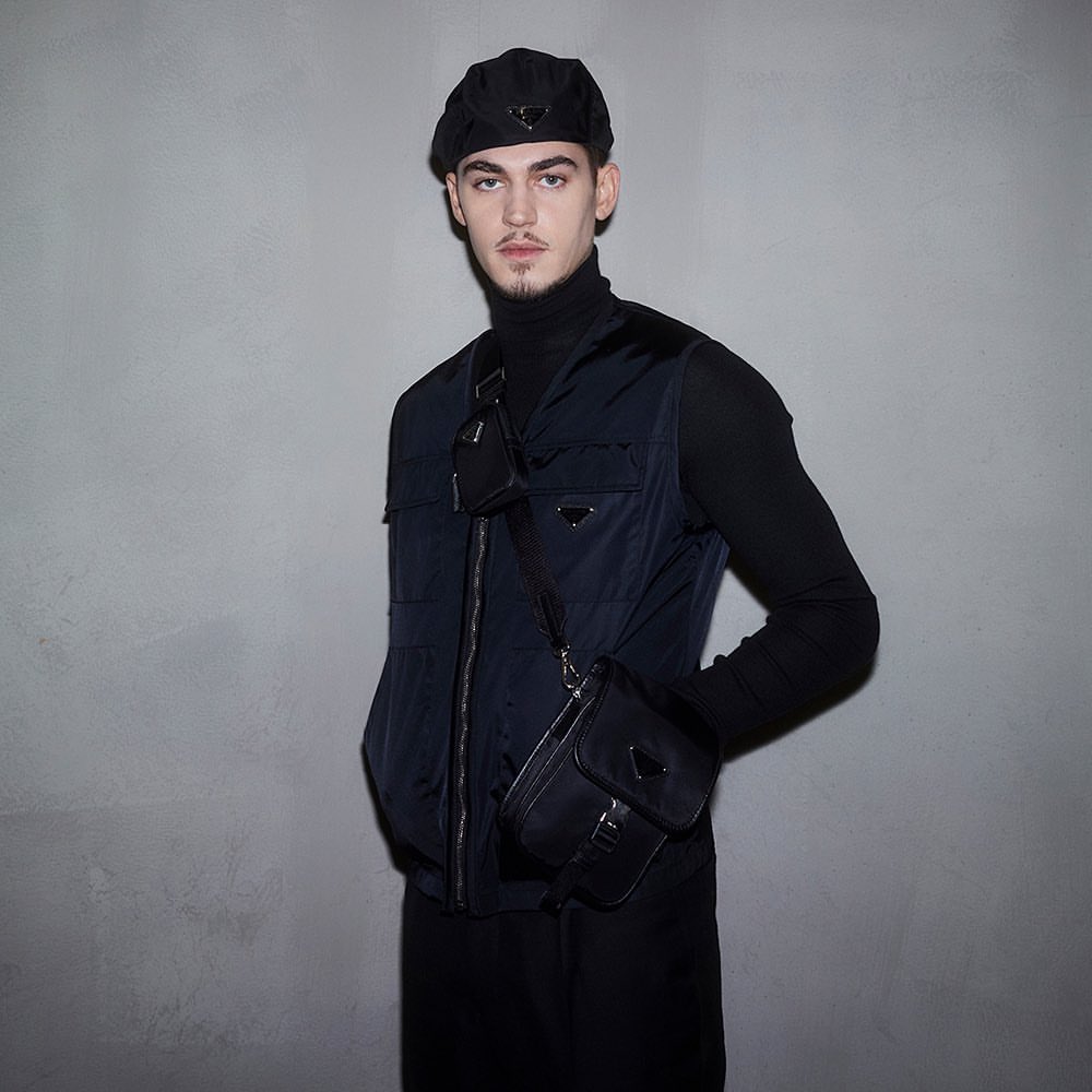 Portrait of a young adult male wearing black turtleneck, navy vest, beret, and cross-body bag against a grey studio backdrop.