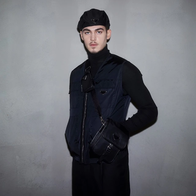 Portrait of a young adult male wearing black turtleneck, navy vest, beret, and cross-body bag against a grey studio backdrop.