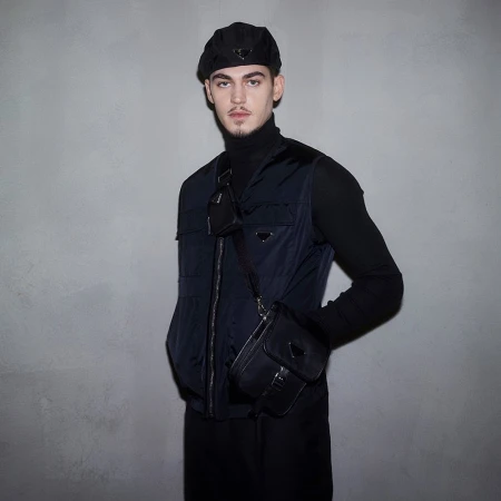 Portrait of a young adult male wearing black turtleneck, navy vest, beret, and cross-body bag against a grey studio backdrop.