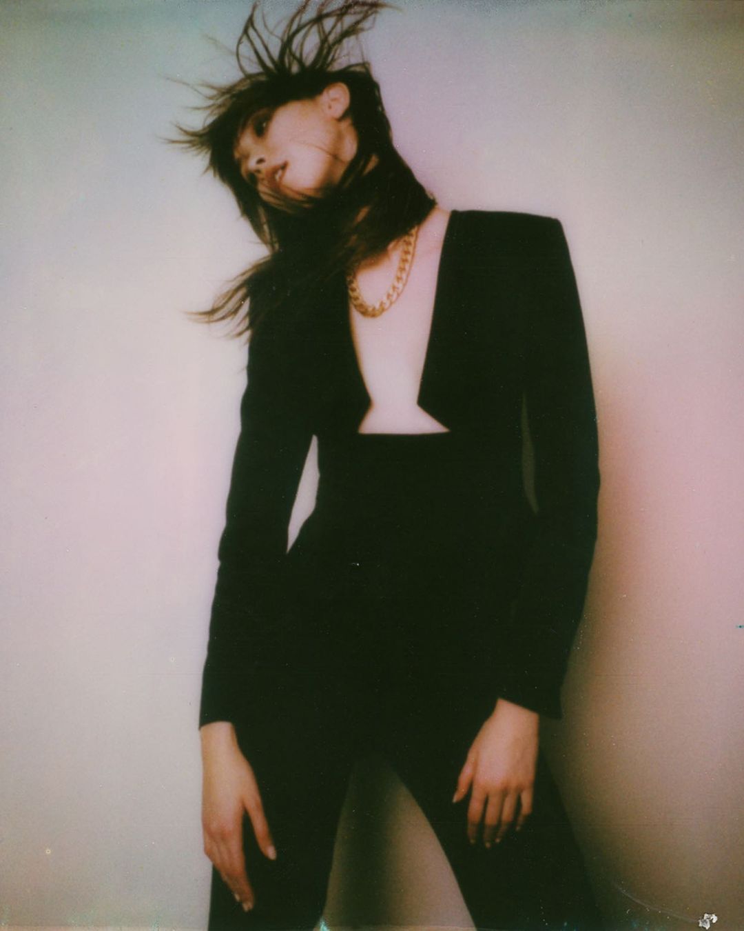 A stylish adult woman with dark hair in a sleek black jumpsuit and gold chain poses against a neutral wall, capturing a modern fashion moment.
