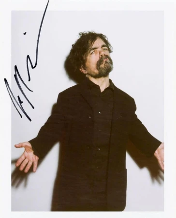 Portrait of Peter Dinklage in a dark jacket against a white background with a signature overlay by Emily Soto