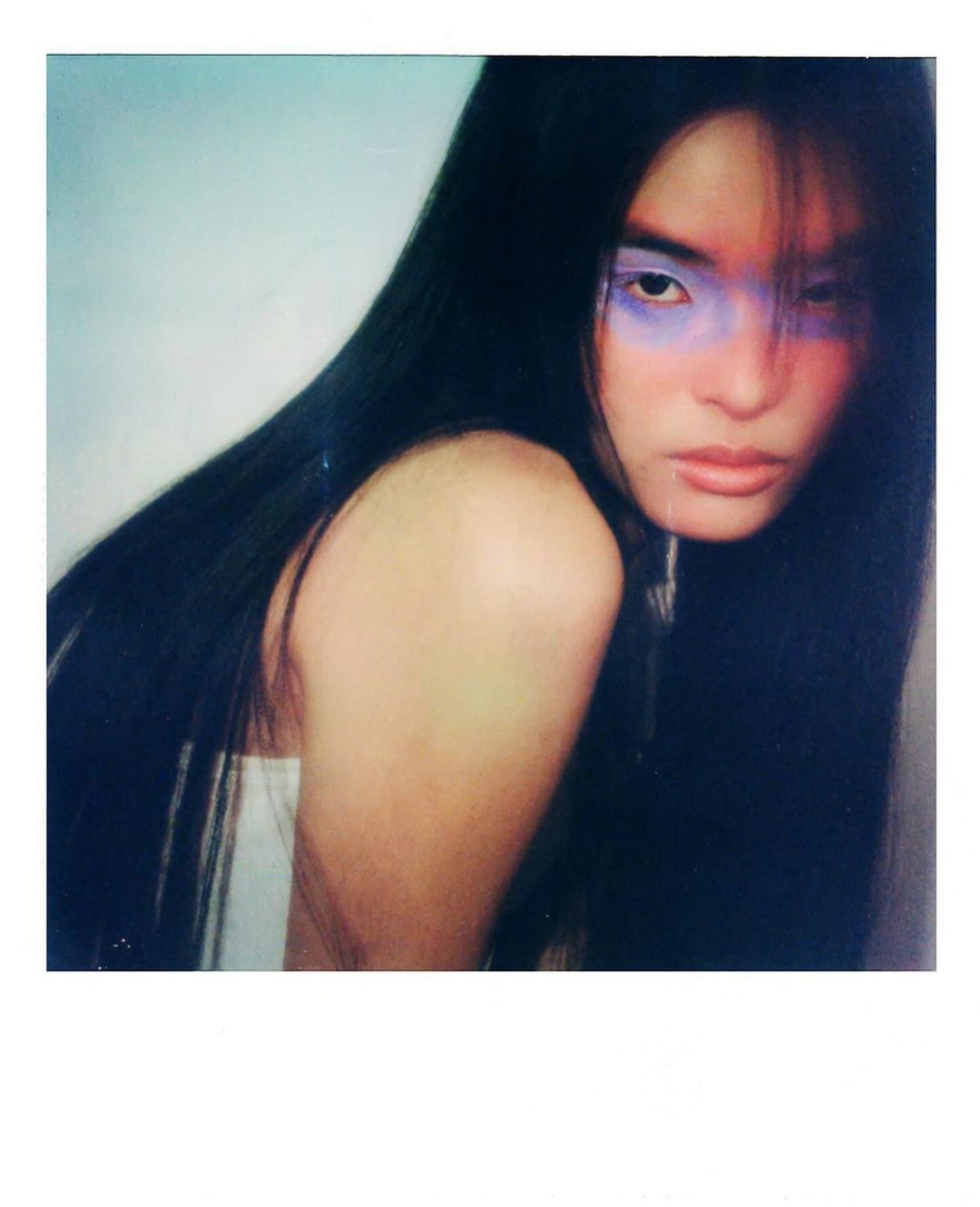 Close-up portrait of an adult female with blue and red eye makeup, dark long hair, and light skin, posing in front of a pale background.
