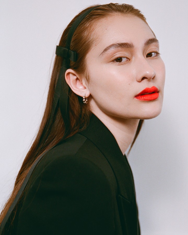 Close-up portrait of a woman with red lipstick, wearing a black blazer and black ribbon headband against a white backdrop.
