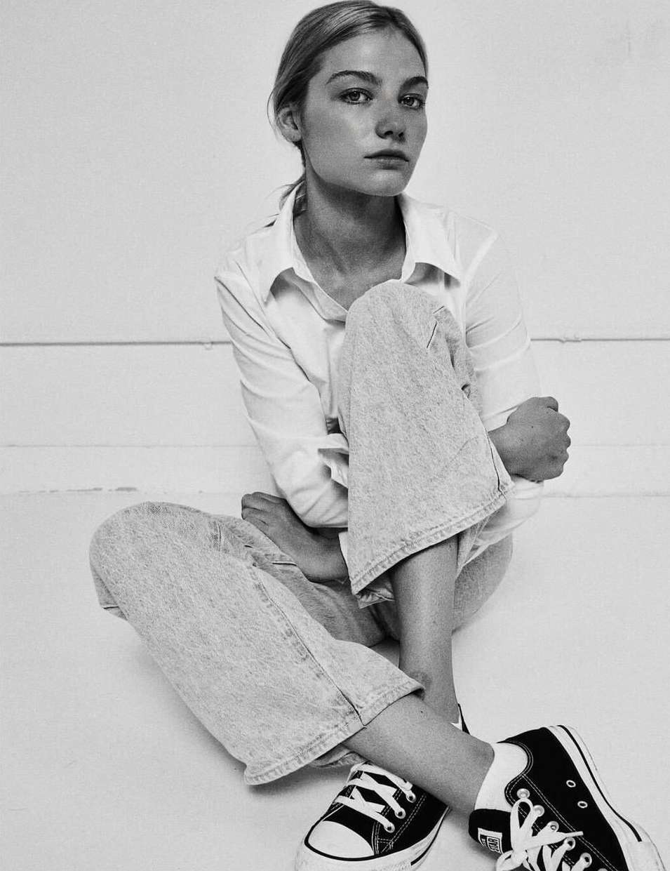 Monochrome portrait of a young woman sitting on the floor wearing a white button-down shirt, light jeans, and sneakers.
