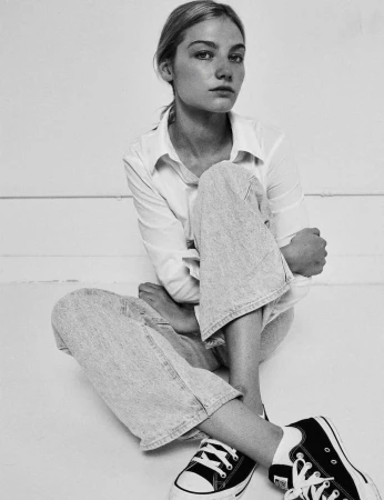 Monochrome portrait of a young woman sitting on the floor wearing a white button-down shirt, light jeans, and sneakers.