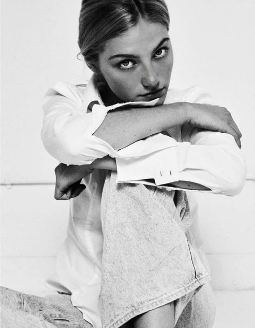 Black and white portrait of a fair-skinned woman with blonde hair in a white button-down shirt and grey jeans, arms crossed over her knee.