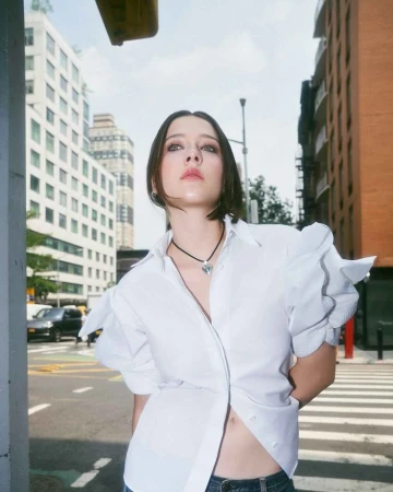 A woman wearing a white buttoned shirt and necklace posing against an outdoor city street background in New York.