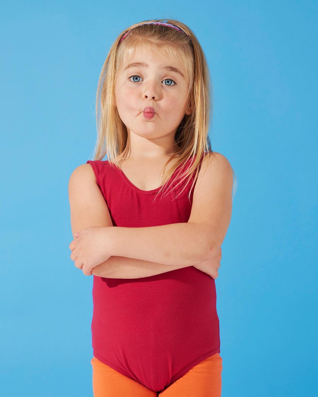 A young girl with fair hair poses with arms crossed, wearing a red leotard and orange shorts against a blue background.