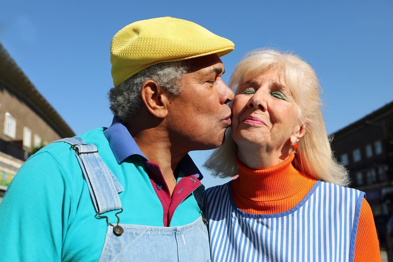 A biracial elderly couple share a kiss outdoors under a sunny sky, with the man in overalls and the woman in a striped apron.