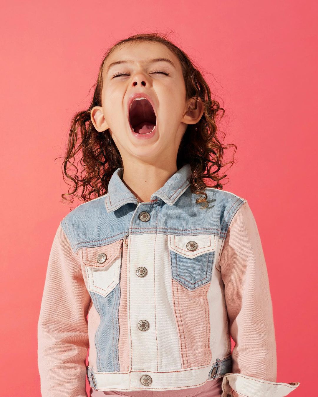 Close-up shot of a young girl yawning with a wide-open mouth. She is wearing a denim jacket with pink and white color blocking.
