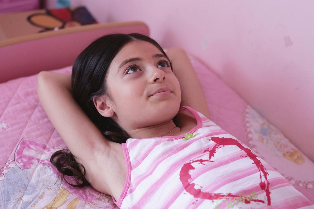 Girl with dark hair lying on a pink bed with hands behind her head, looking up and to the right. Girl wearing a striped top.