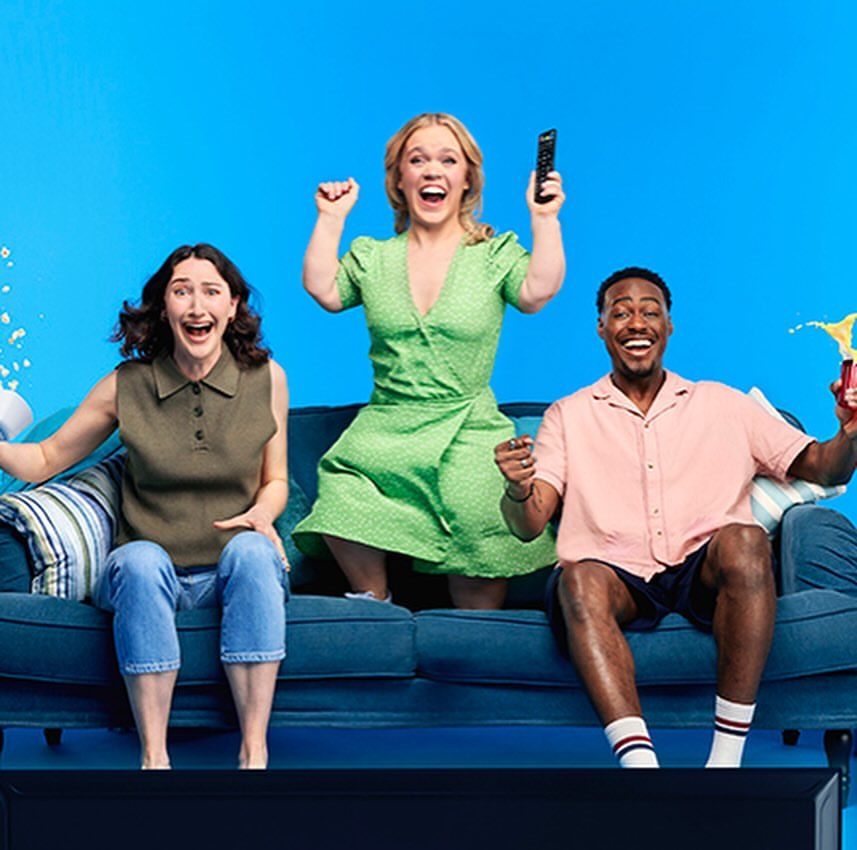 Three excited people celebrate while sitting on a blue sofa in front of a television against a light blue backdrop.
