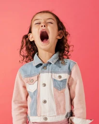 Child yawning with eyes closed, wearing a pink and blue denim jacket against a solid pink background.