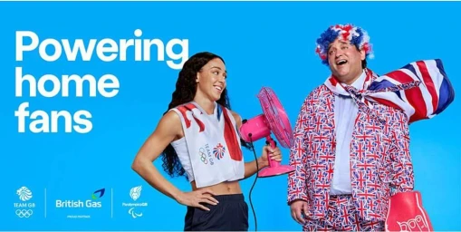 Woman in athletic top holding a pink fan next to a man in a Union Jack suit and wig against a blue background
