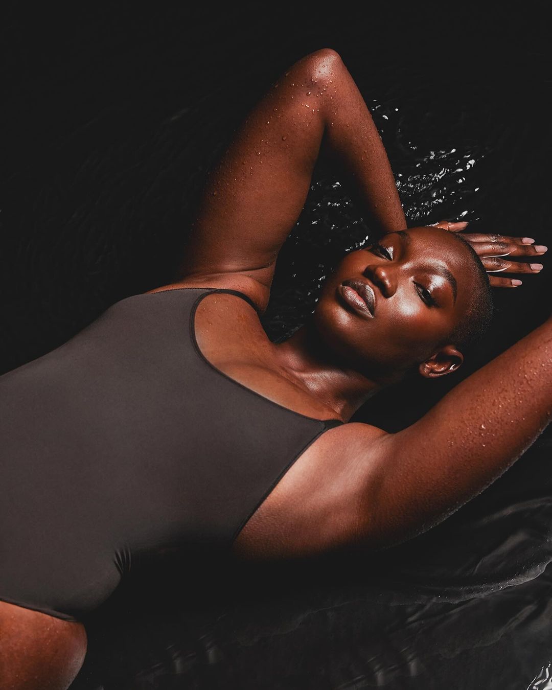 A dark-skinned woman with short hair lying in water, wearing a dark gray one-piece swimsuit. She has her arms above her head.