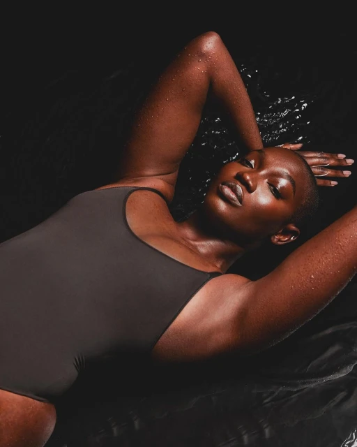 A dark-skinned woman with short hair lying in water, wearing a dark gray one-piece swimsuit. She has her arms above her head.