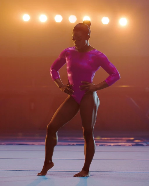 Full-length portrait of an athletic woman in a pink leotard posing confidently on a blue floor with bright studio lighting.