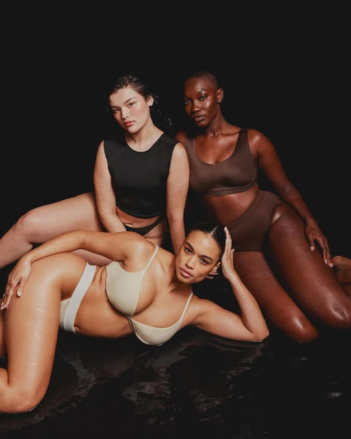 Three adult women pose together wearing neutral color undergarments, lying in a dark environment with low-key lighting.