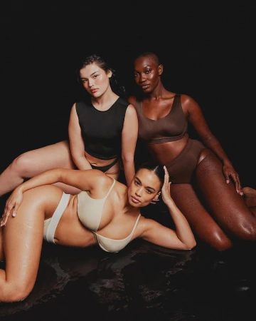 Three adult women pose together wearing neutral color undergarments, lying in a dark environment with low-key lighting.