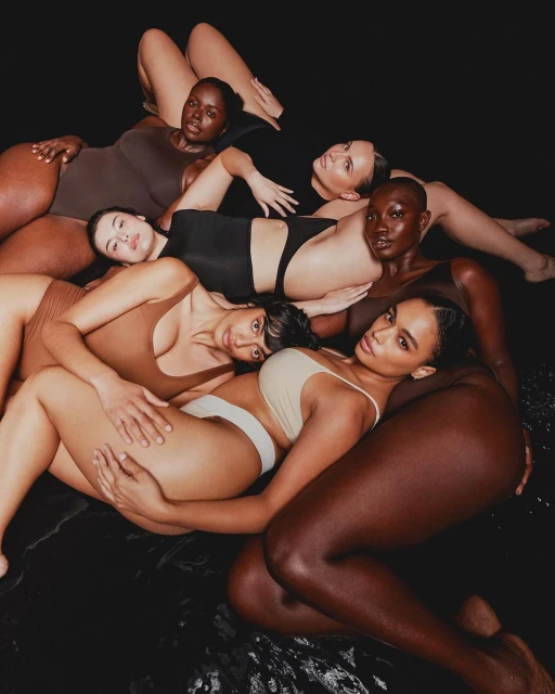 Seven diverse women with varied skin tones pose in a dark, wet studio, wearing neutral-toned underwear and swimsuits, with a focus on body diversity.