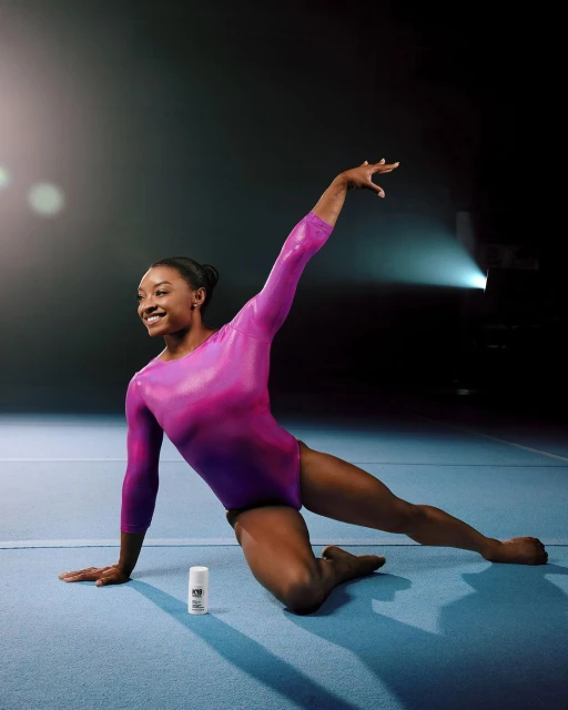 Gymnast Simone Biles in a dynamic pose, wearing a long-sleeved pink leotard on a blue mat next to deodorant, studio lighting and dark background.