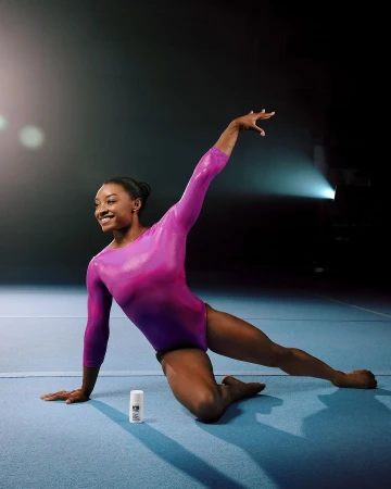 Gymnast Simone Biles in a dynamic pose, wearing a long-sleeved pink leotard on a blue mat next to deodorant, studio lighting and dark background.