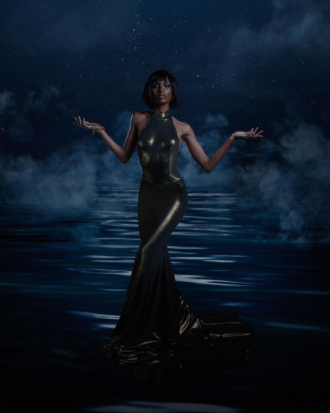 Elegant Black woman in a golden gown, arms extended, posing against a night sky backdrop with a body of water and fog.