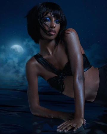 Elegant woman with dark skin and blue eyeshadow poses with black top in dim lighting against a blue background with faint moon.