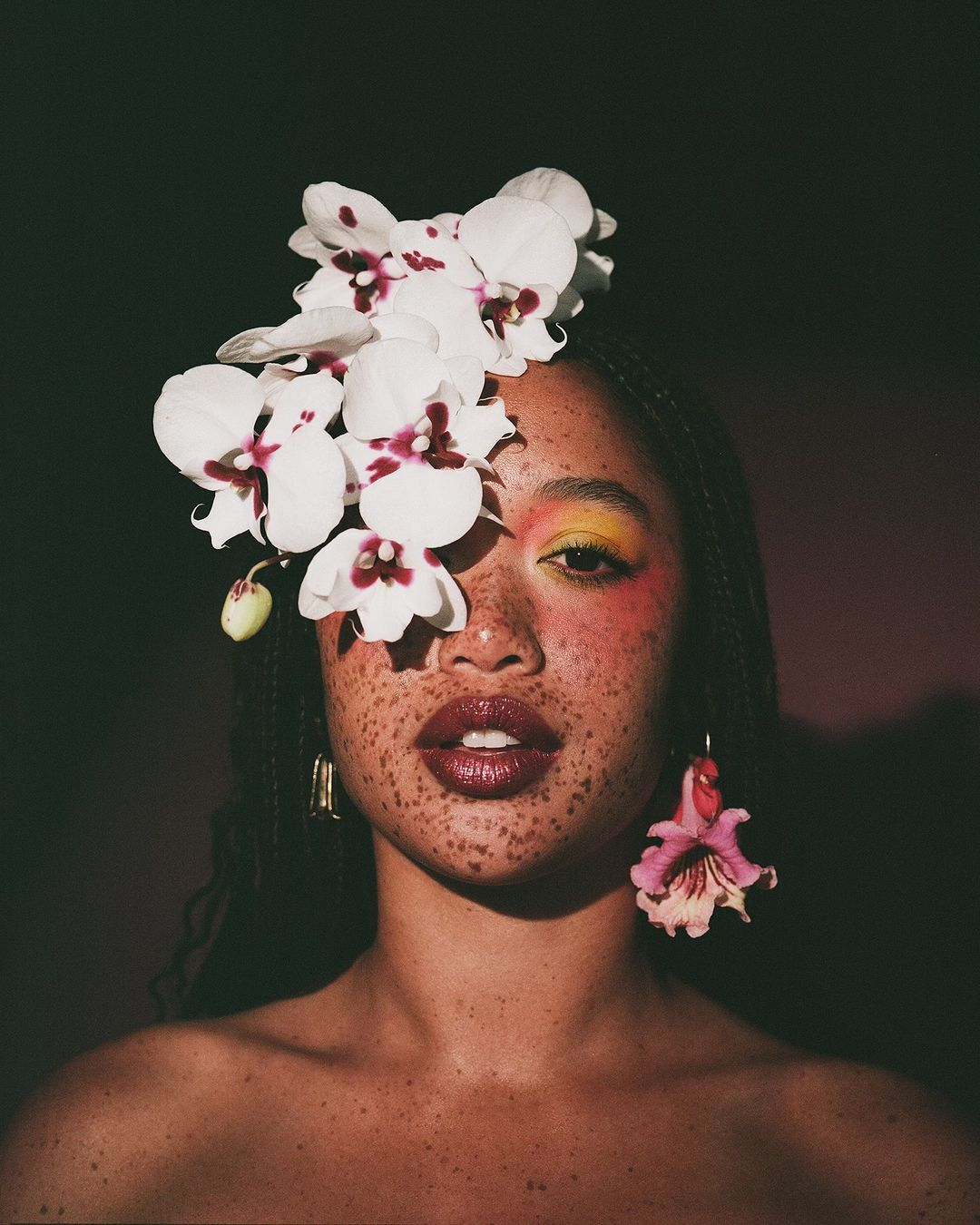 Close-up studio shot of a woman with freckles and colorful makeup, wearing a headdress of white orchids and a matching earring.
