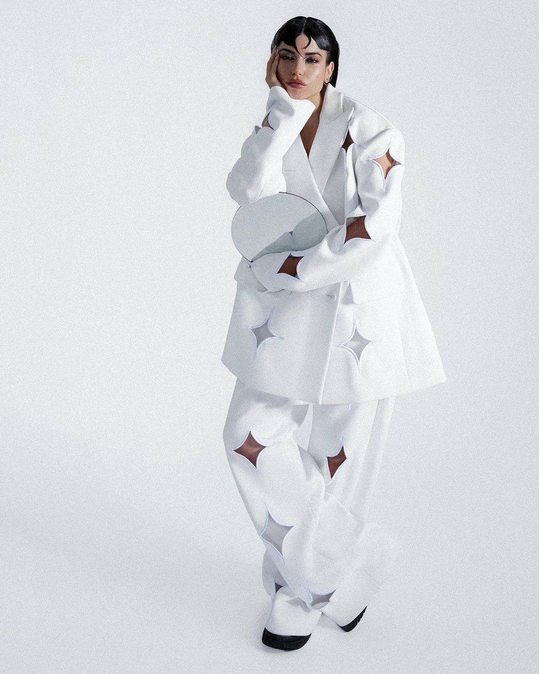 Fashion model poses in studio wearing a white suit with star cutouts, holding a round mirror. Soft, diffused lighting creates a dreamy effect.