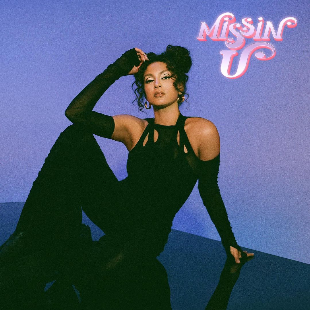 A woman poses in a black catsuit against a blue background with the text “Missin U” visible; fashion and beauty concept.