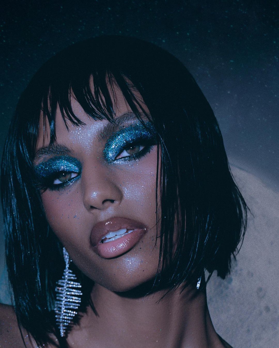 Close-up portrait of an adult female with short black hair, glittery blue eyeshadow, wearing leaf-shaped crystal earrings, in studio.
