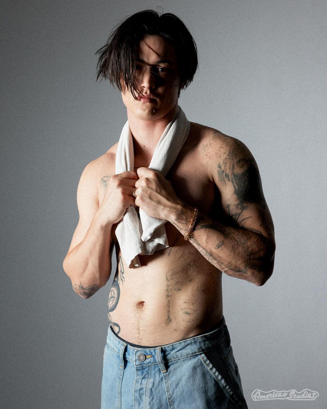 Shirtless man with tattoos wearing light wash denim jeans and holding a white towel draped around his neck in a studio portrait.