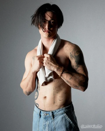 Shirtless man with tattoos wearing light wash denim jeans and holding a white towel draped around his neck in a studio portrait.