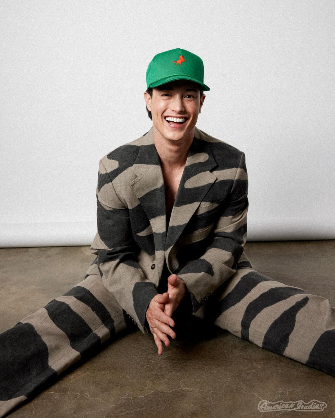 A smiling Asian man sits on the floor wearing a green baseball cap with an orange logo and a grey-and-black zebra-striped suit.