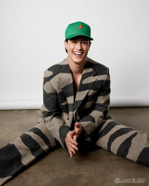A smiling Asian man sits on the floor wearing a green baseball cap with an orange logo and a grey-and-black zebra-striped suit.