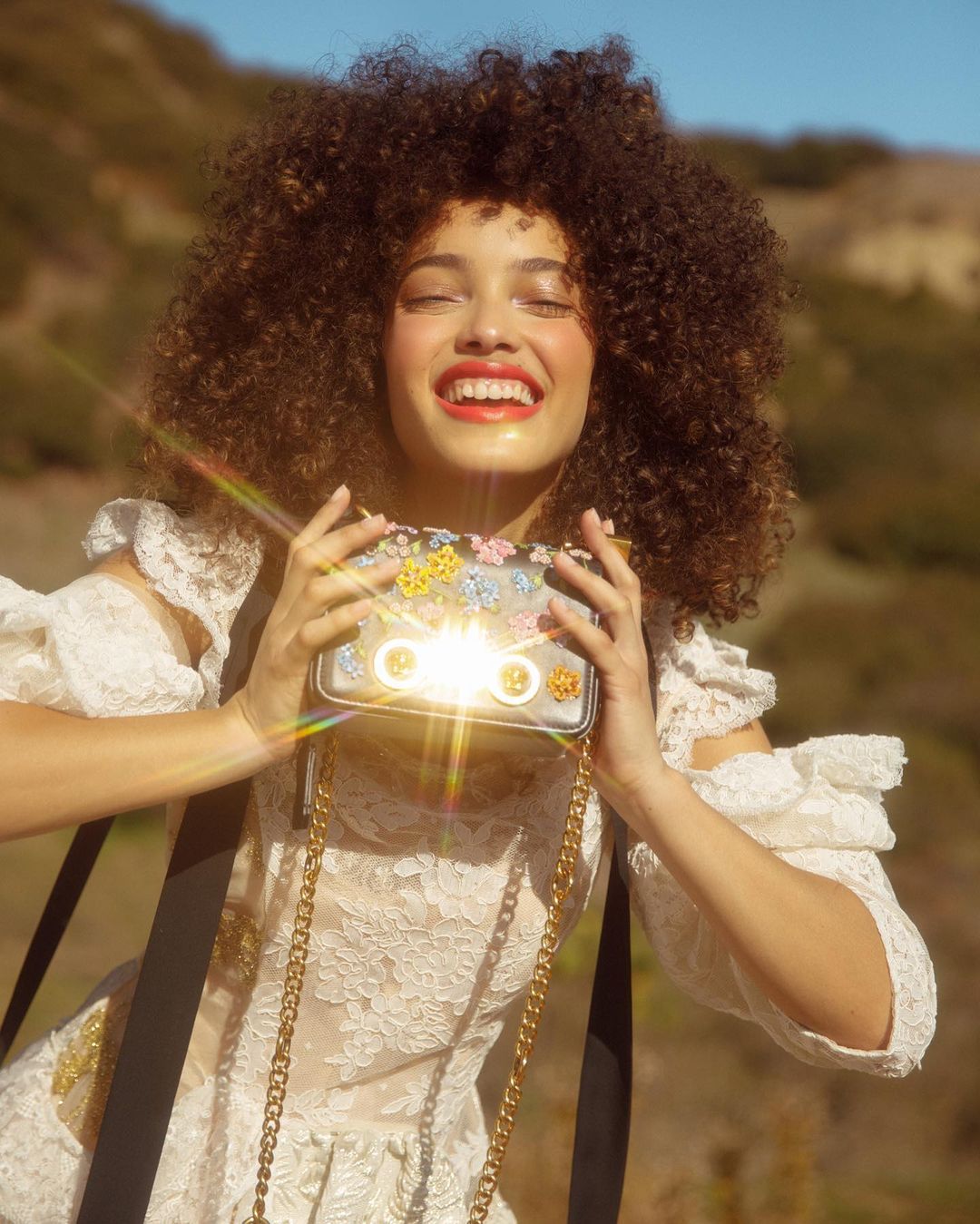 A happy adult female smiles while holding a small floral purse outdoors in a medium shot with a blurred natural background.