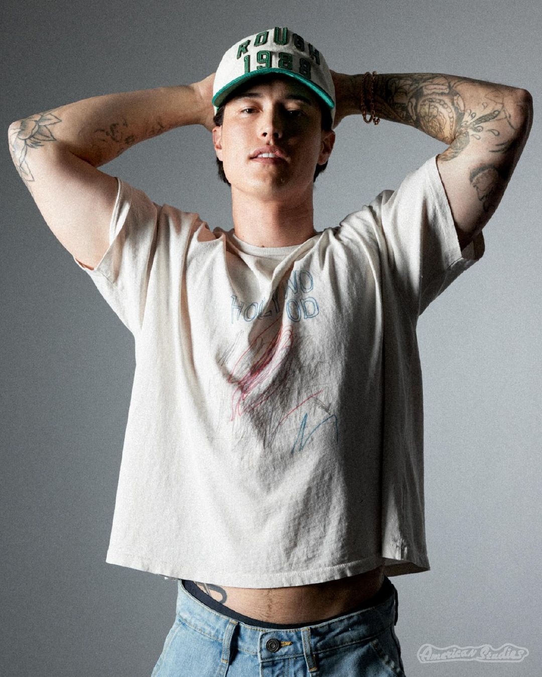 Young man wearing a white baseball cap and graphic t-shirt with hands behind his head in a studio portrait by Emilynn Rose