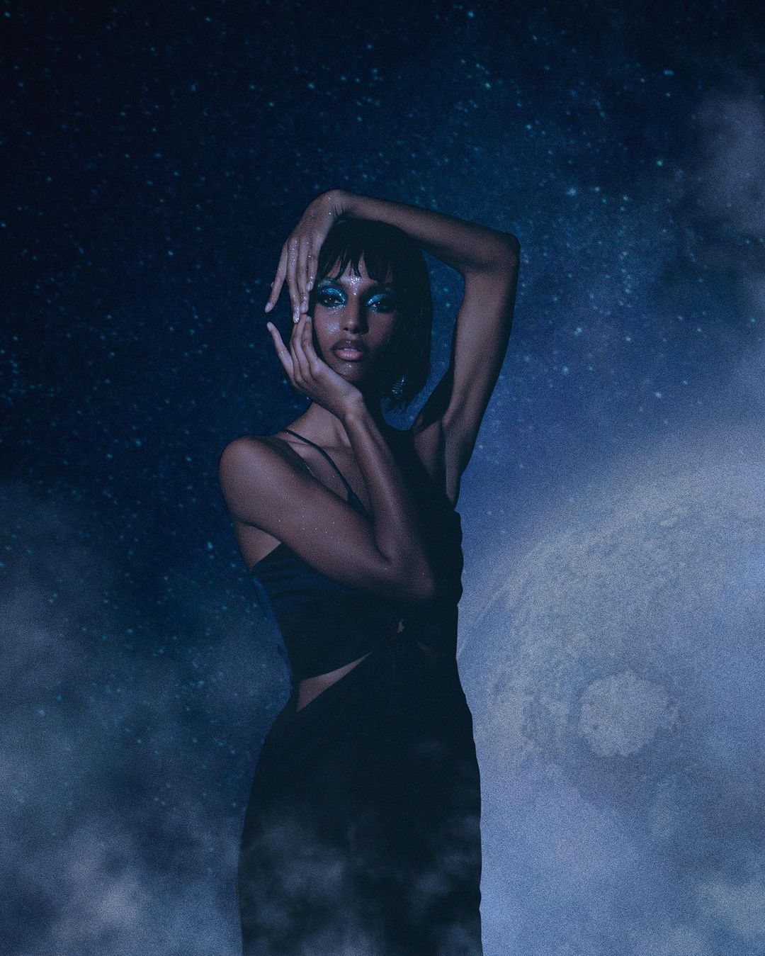 Portrait of a young woman with blue eyeshadow, posing against a dreamy background of stars and a moon, wearing a black dress.