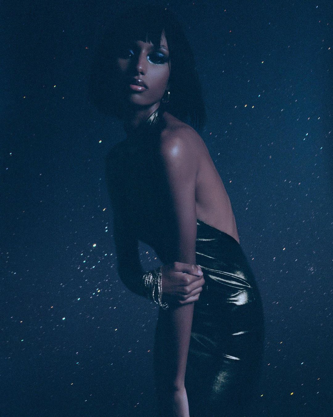 Glamorous portrait of an African American woman in a shiny dress. She poses against a dark blue background with star-like specks of light.