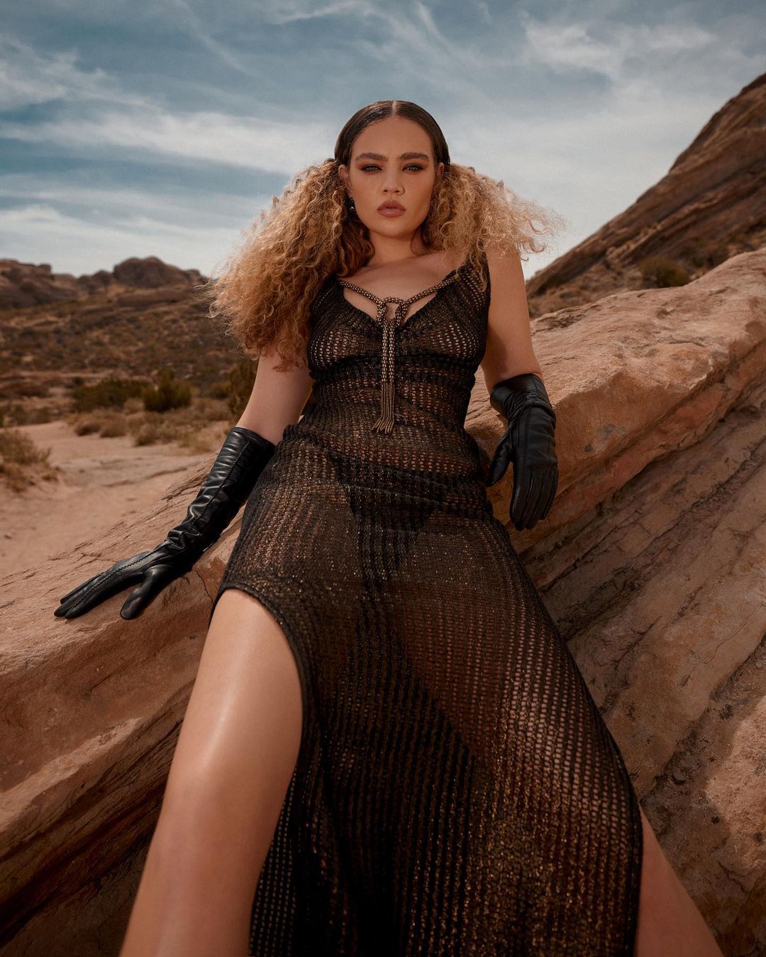 Woman with curly blonde hair poses in a sheer black dress with a thigh-high slit in a desert landscape with a rock formation backdrop.