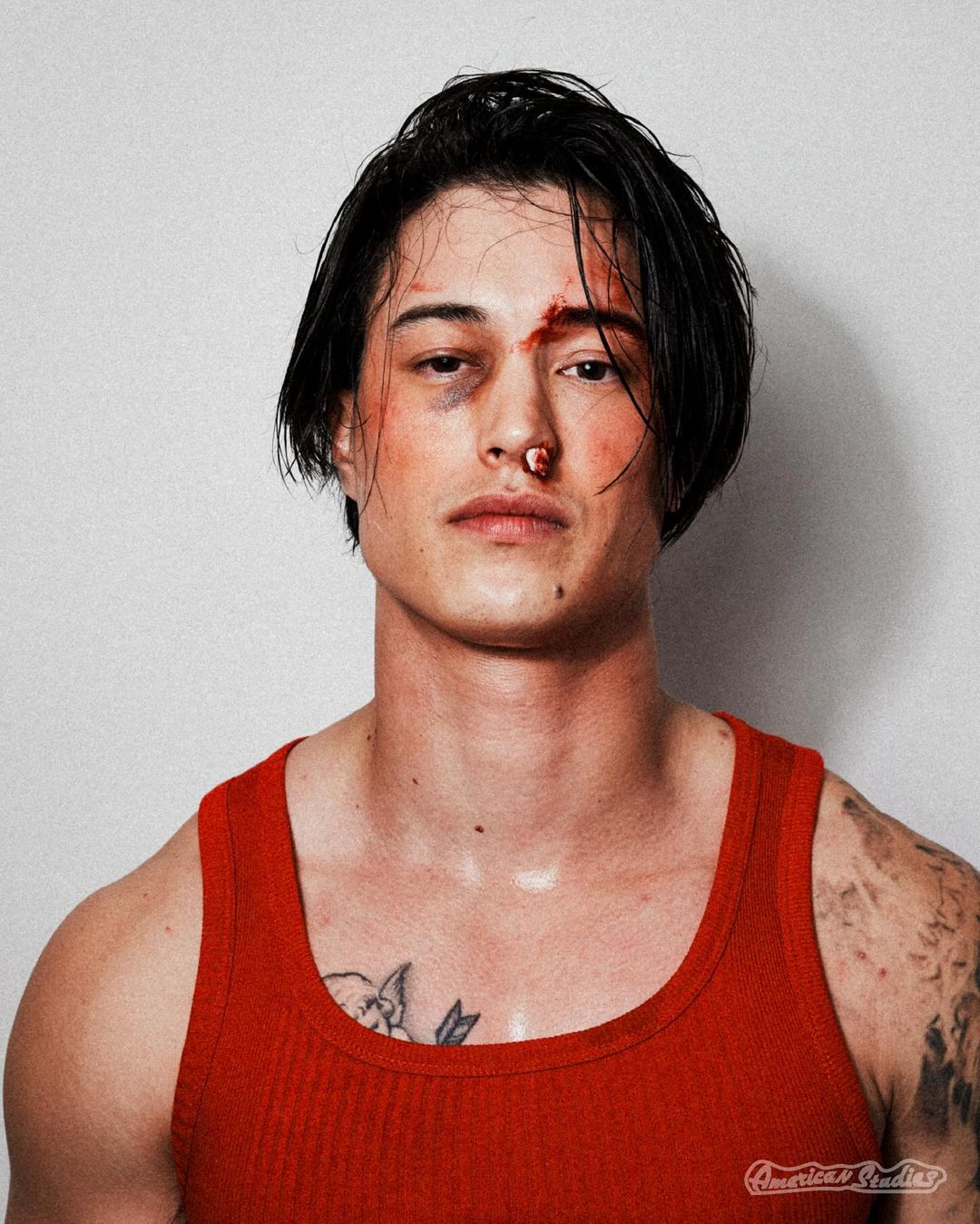 Close-up portrait of an adult male with an injury on his face, wearing a red tank top against a neutral background indoors.