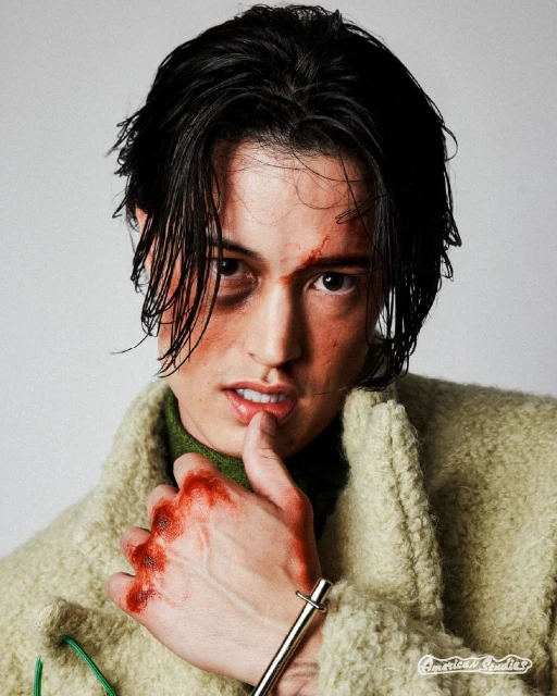 Portrait of an adult male with visible injuries, wearing a wool coat, holding a hand near his mouth in a light studio setting.