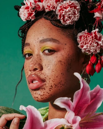 Close-up portrait of a freckled woman with floral headpiece and yellow eyeshadow, holding pink lily, against a teal backdrop.